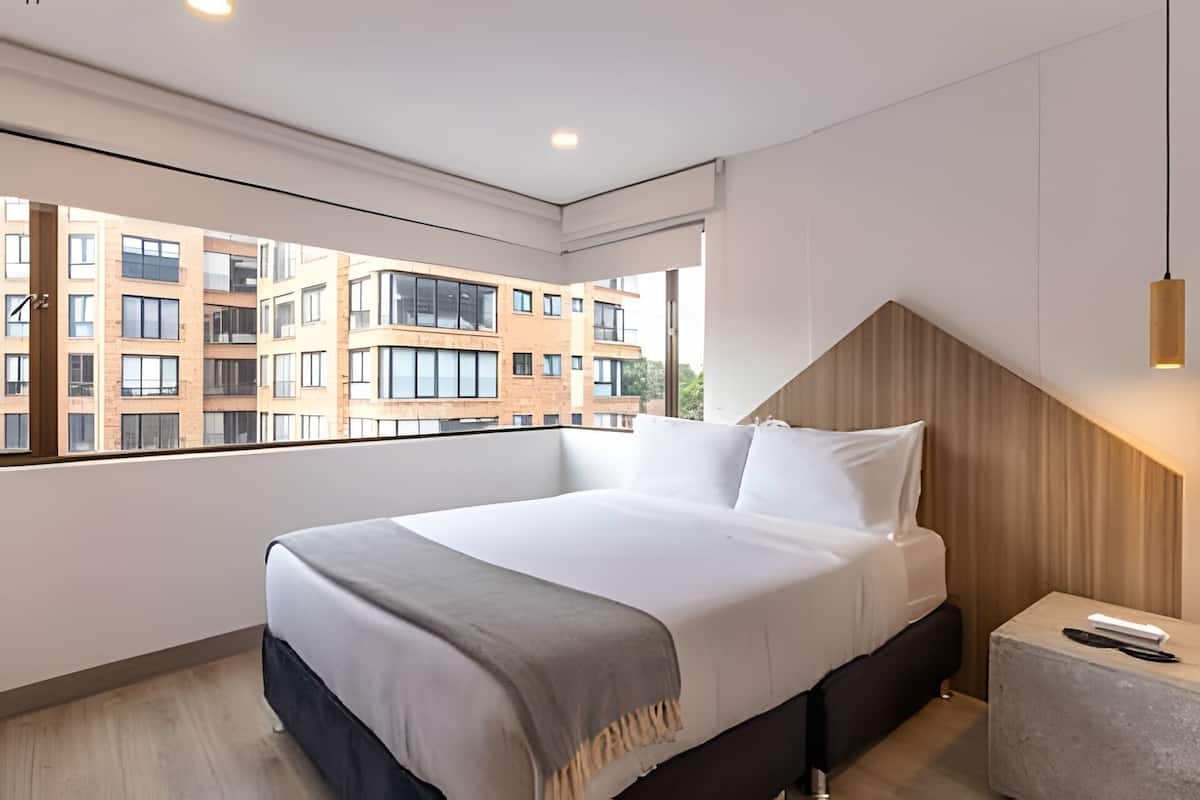 Une chambre d’hôtel moderne avec un grand lit, une tête de lit en bois, une table de chevet et une vue sur un immeuble par la fenêtre.