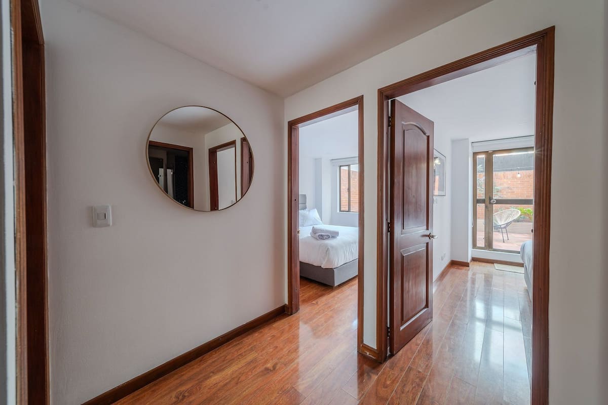 Un couloir au plancher en bois, un grand miroir rond accroché au mur, et une chambre à coucher visible à travers une porte ouverte.