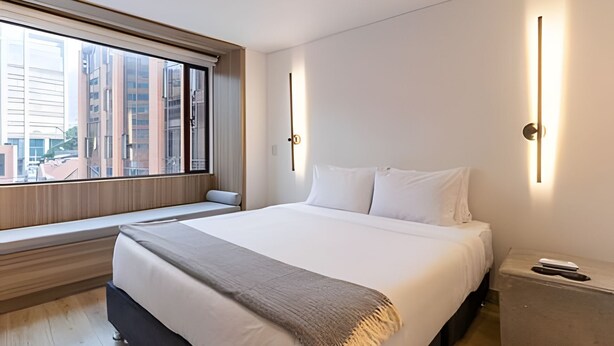A modern hotel room with a large bed, a bedside table, a window with a view of buildings, and a wall-mounted light fixture. - 10.73 Flats x Ahead (Bogotá)