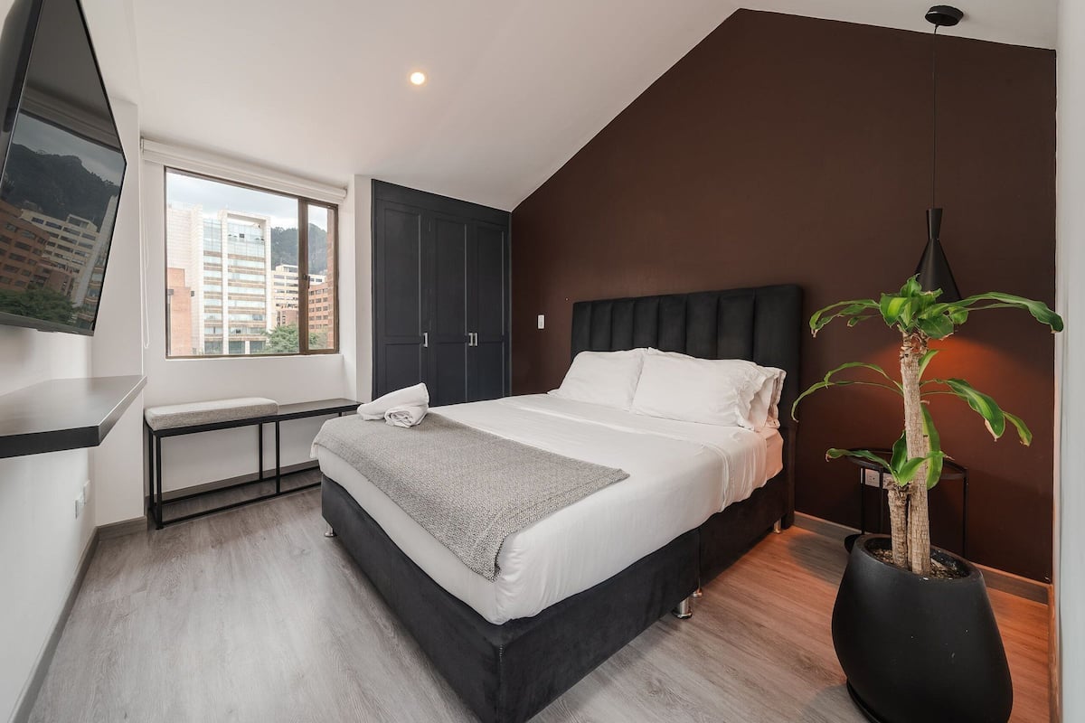 Une chambre à coucher moderne avec un lit, un banc, une plante en pot et une vue sur les bâtiments à travers une fenêtre.