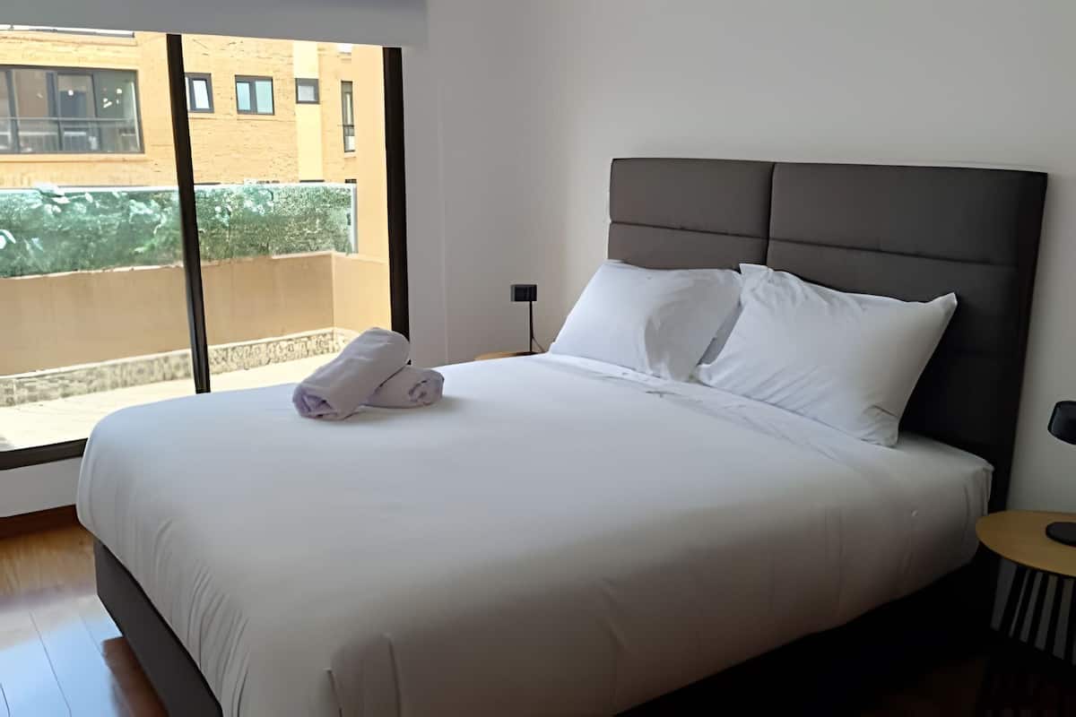 Un lit bien fait, avec des draps blancs, une tête de lit grise et une paire de serviettes pliées posées sur le lit.