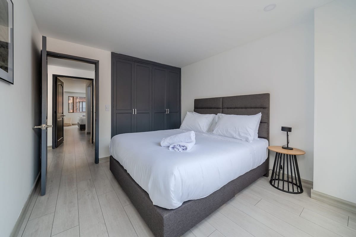 Un lit bien fait, avec du linge de lit blanc, une petite table ronde avec une lampe, et une armoire sombre dans une chambre d’hôtel moderne.