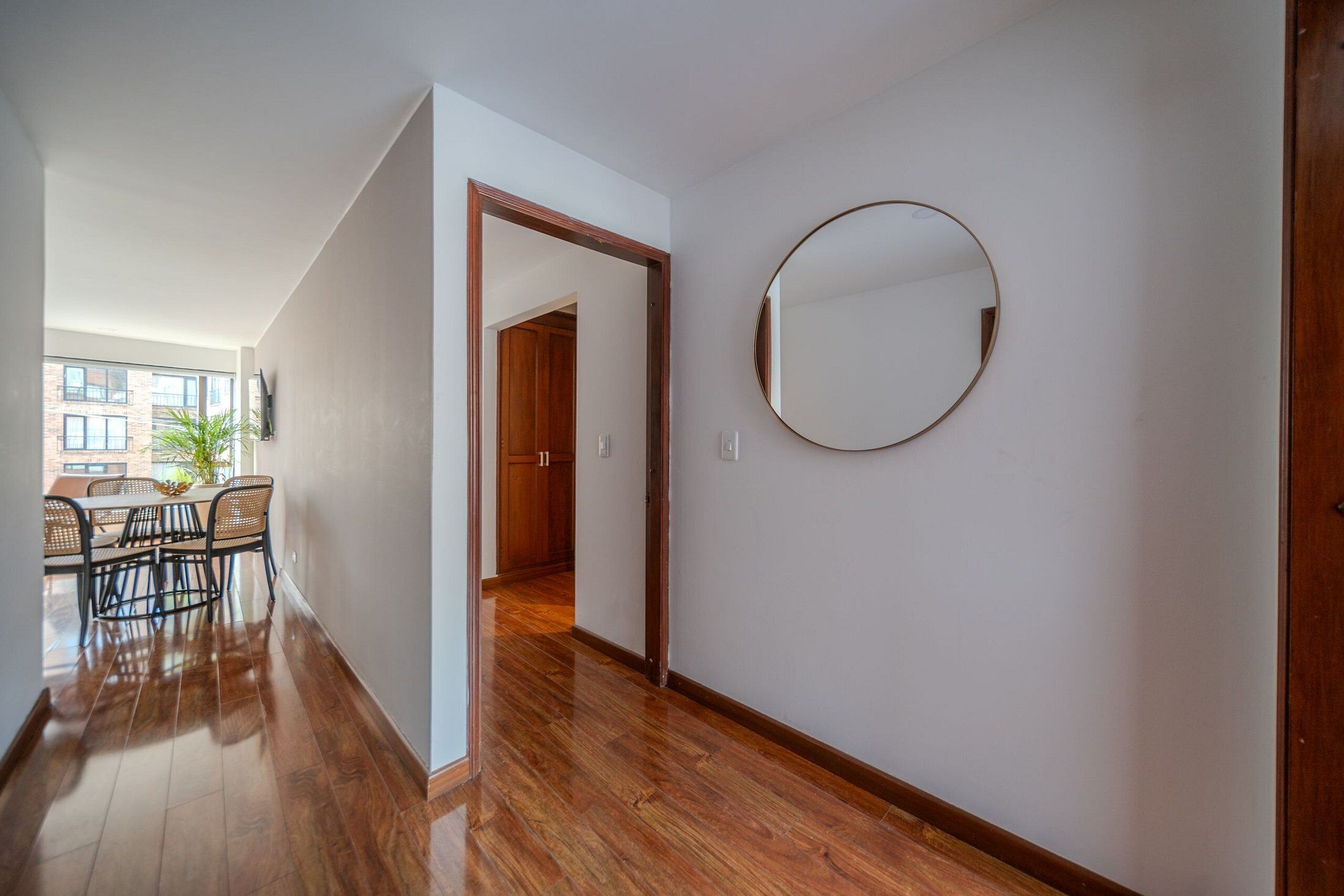 A hallway with wooden flooring, a round mirror on the wall, and a dining area with chairs and a table.