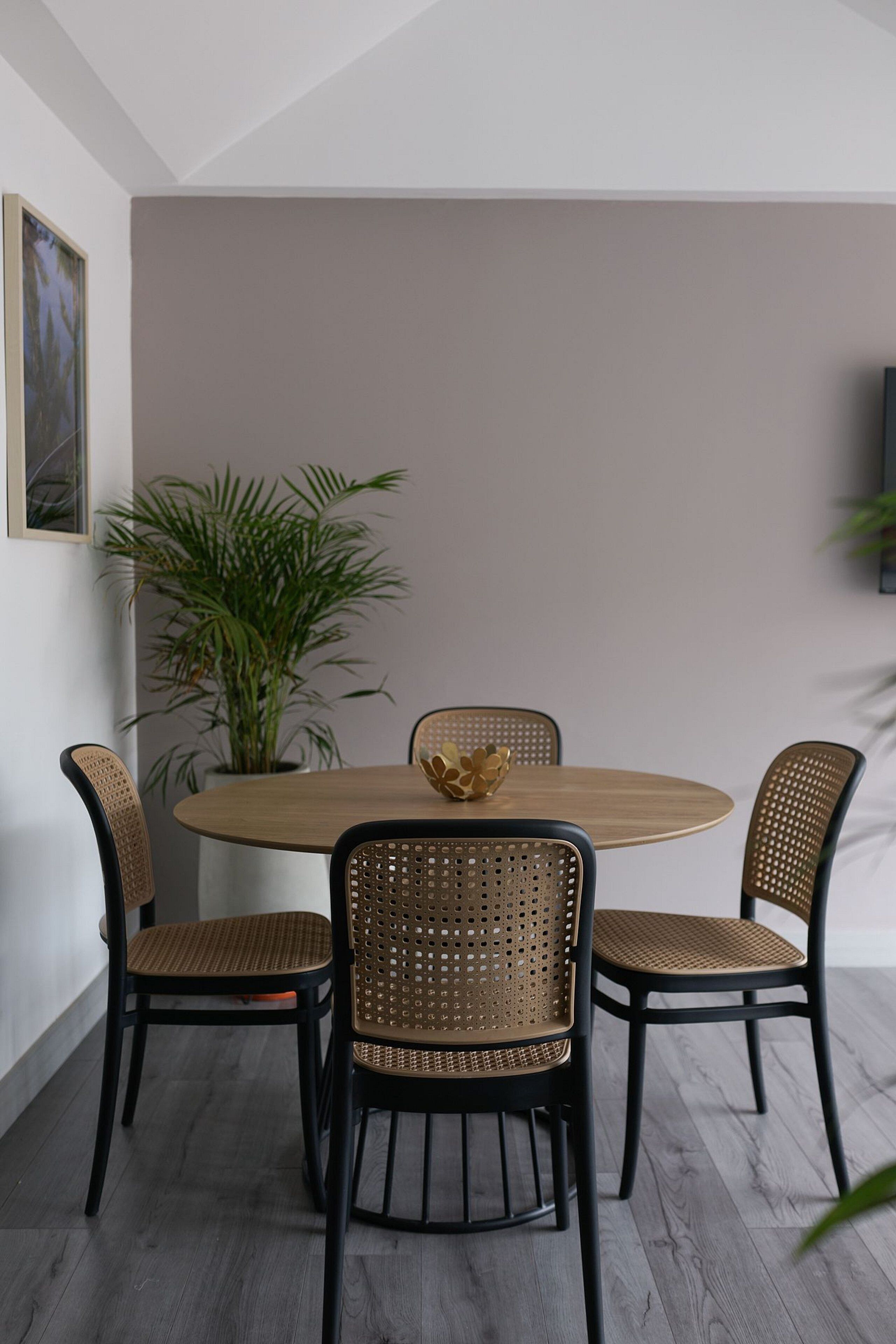 Uma área de refeições com uma mesa redonda, quatro cadeiras de vime, um vaso de planta e uma televisão na parede.