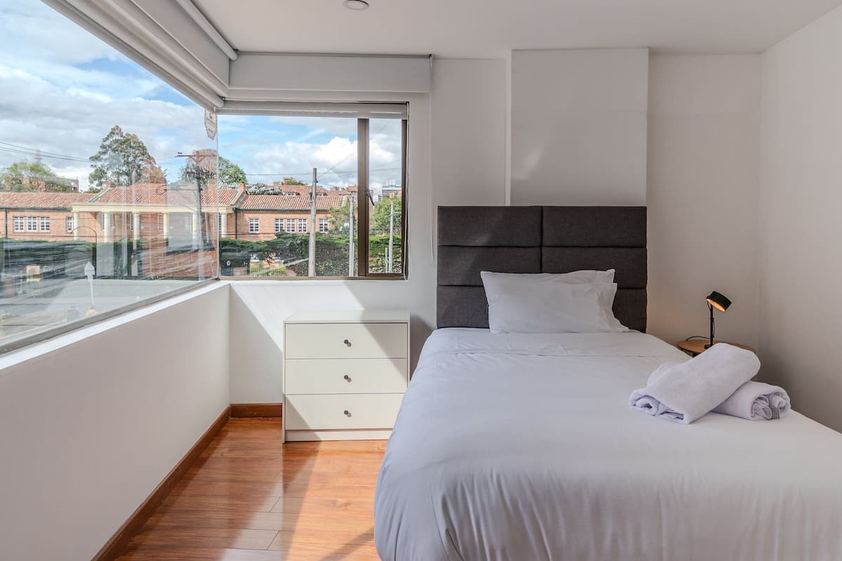 Une chambre avec une grande fenêtre, un lit recouvert de draps blancs, une lampe de chevet et une petite commode blanche.