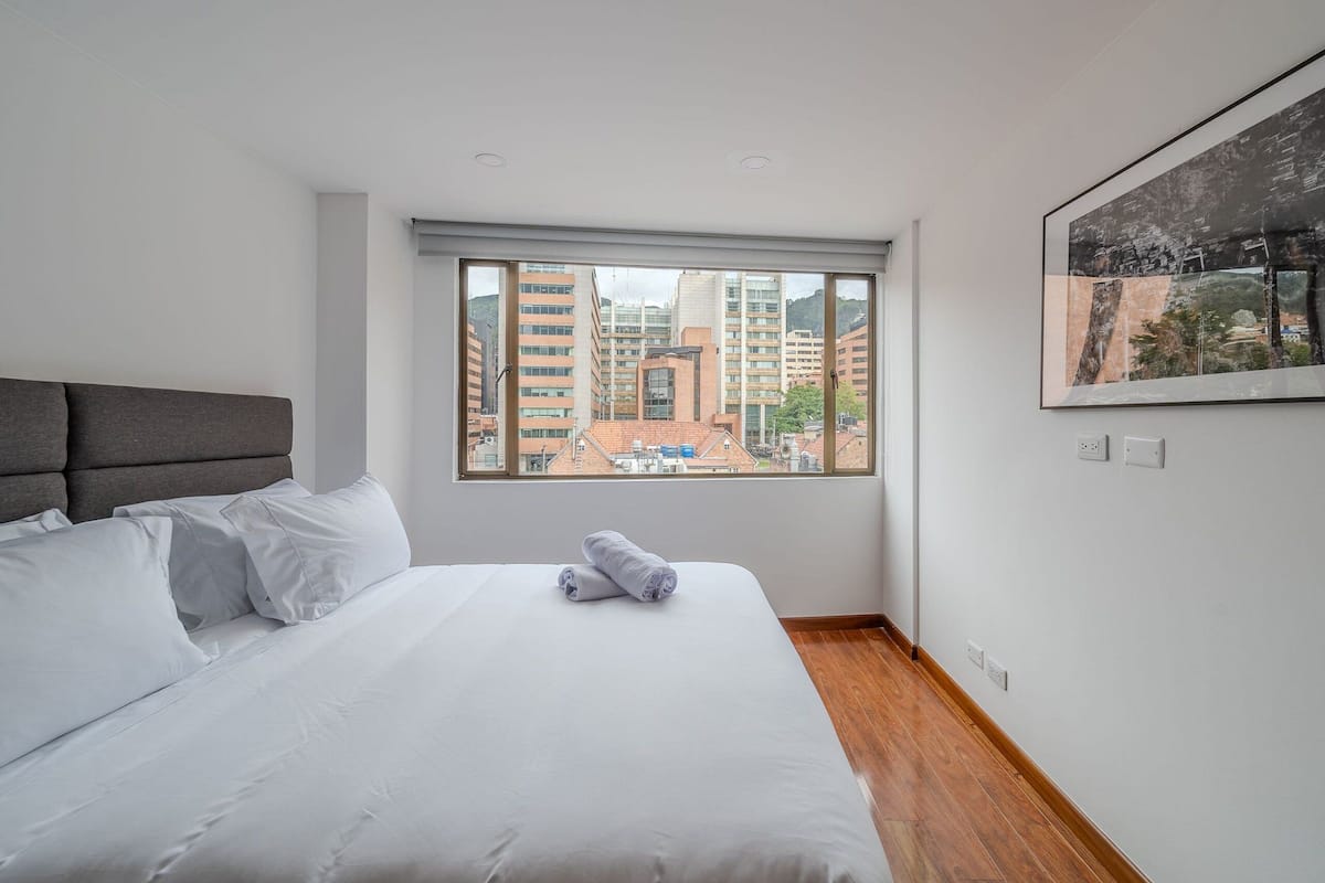 Une chambre d’hôtel avec un grand lit, une tête de lit, une fenêtre donnant sur la ville et un tableau encadré au mur.