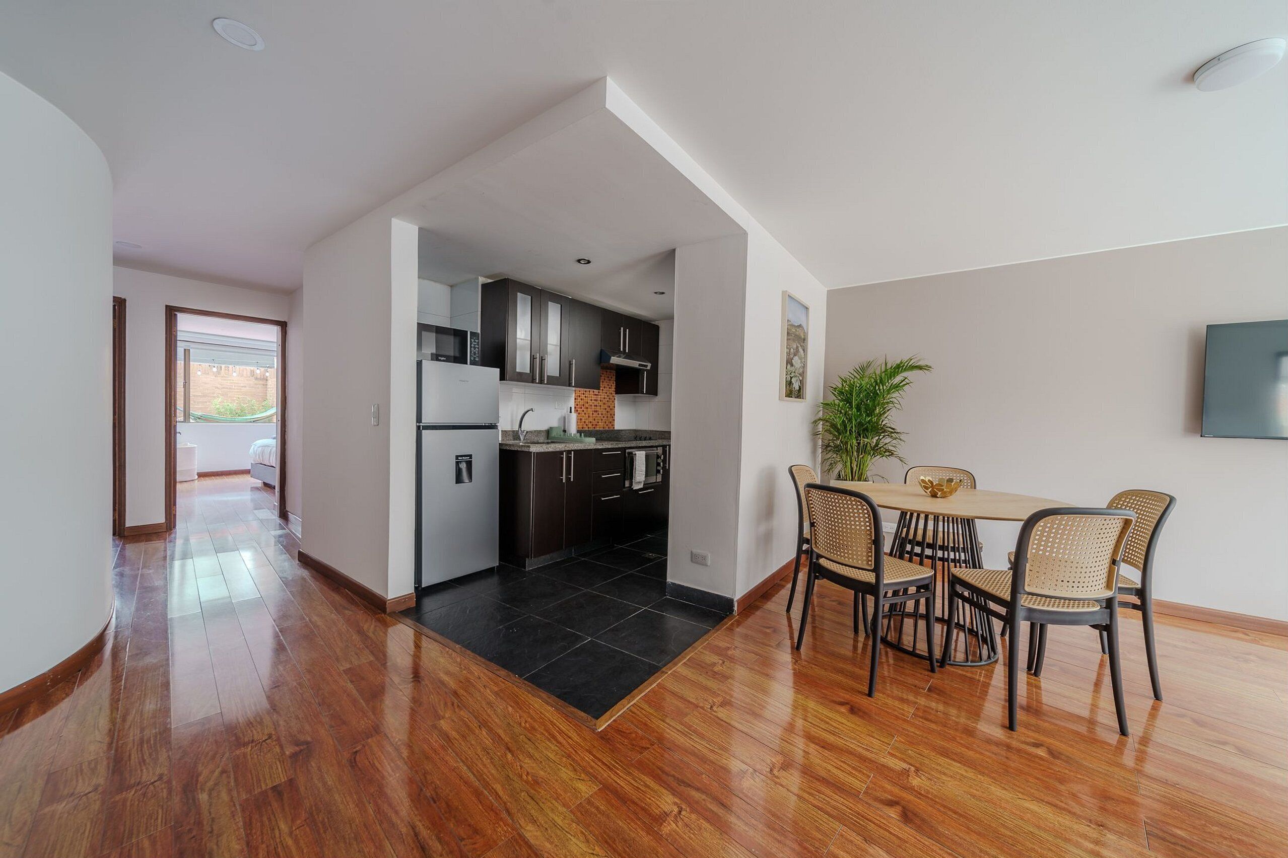 A modern kitchen with dark cabinets, a central island, and a dining area with wicker chairs and a table.