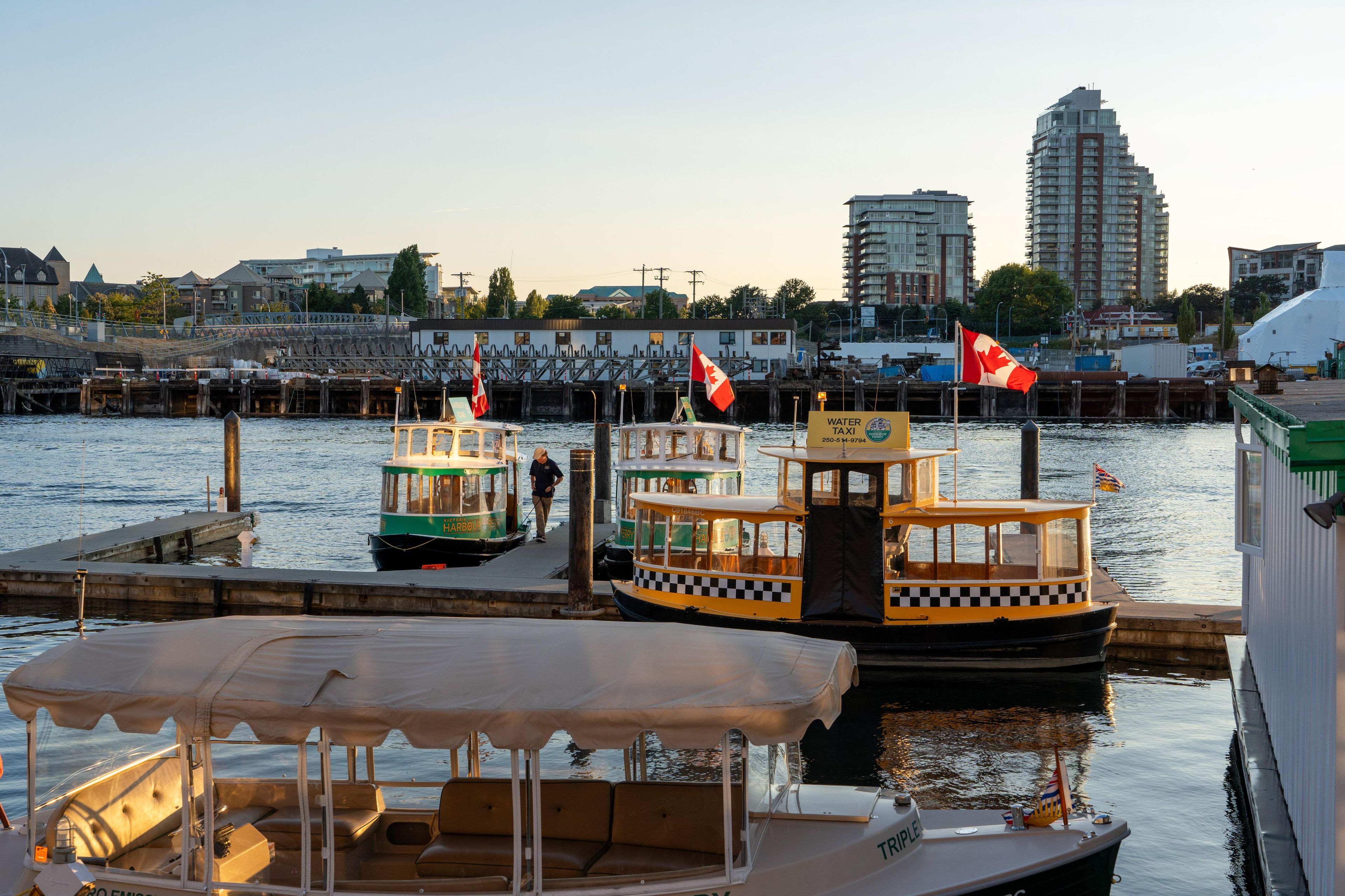 A waterfront marina with several boats docked, a bridge in the background, and modern buildings.