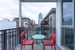 Un balcon avec deux chaises rouges et une petite table ronde, donnant sur une rue bordée d’immeubles et où l’on aperçoit, au loin, un grand gratte-ciel.