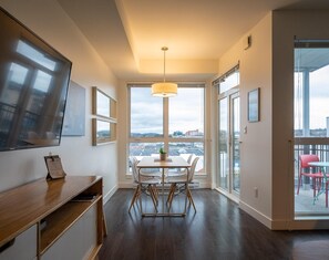 A modern dining area with a table and chairs, a pendant light, and a view of the city through large windows. - Bright and Trendy Heritage Suite in Old Town (Victoria Central British Columbia Provincial Government)