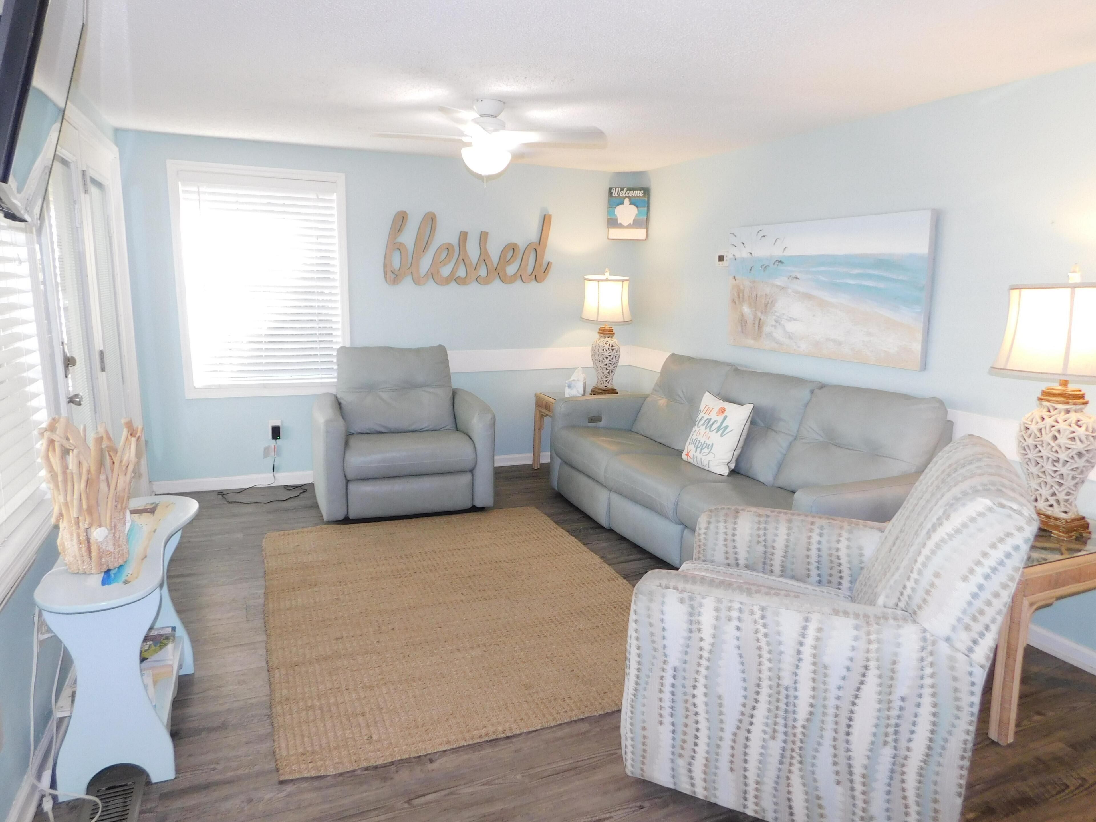 A living room with a grey sofa, a grey armchair, a small table with a lamp, a rug, and a wall with a 'blessed' sign.