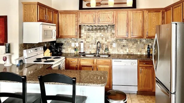 A kitchen with wooden cabinets, granite countertops, a stainless steel refrigerator, and a central island with chairs.