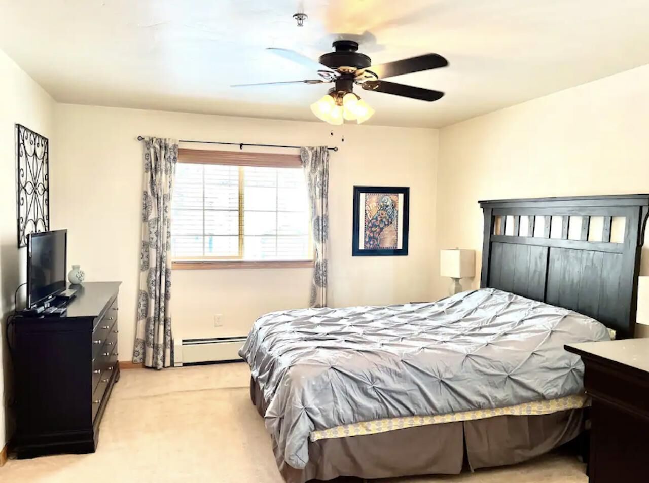 A bedroom with a bed, a ceiling fan, a dresser, a window with curtains, and a framed picture on the wall.