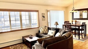 A living room with a sofa, a coffee table, a dining table with chairs, a chandelier, and a large window with blinds.