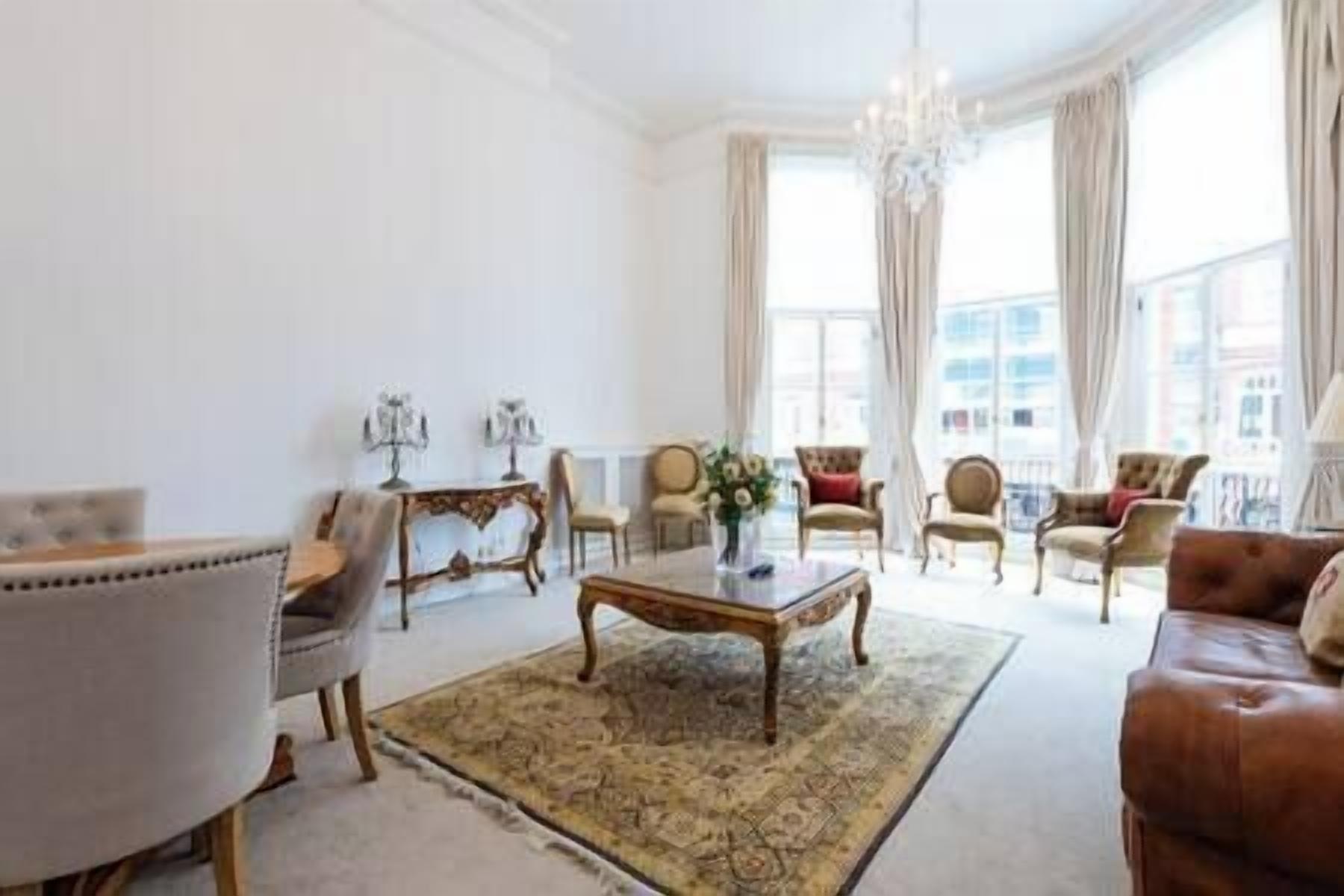 A spacious living room with a large coffee table, several armchairs, and a sofa. There is a chandelier hanging from the ceiling, and large windows with curtains.