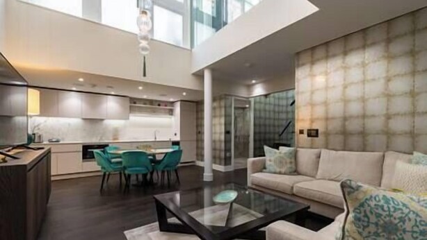 A modern living room with a glass coffee table, a sofa with patterned cushions, and a dining area with teal chairs.