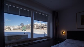 A hotel room with a bed, a nightstand, a wall-mounted lamp, and a window with a view of a snowy landscape and a parking lot. - Panorama App 1B Zell am See (Zell am See)