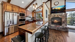 A modern kitchen with stainless steel appliances, wooden cabinets, a large island with stools, and a stone fireplace. - Top of Kingsbury View Home 7BR,4BA,Spa, 5 To Ski (Stateline)