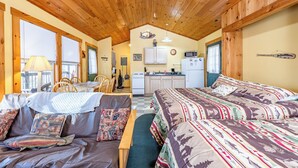 A cozy cabin interior with a wooden ceiling, a dining area, a kitchenette, and a large bed with patterned bedding.