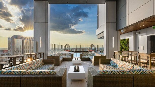 A rooftop lounge with wicker furniture, a central white table, and a view of the city skyline.