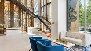 A modern lobby with a reception desk, blue chairs, a staircase, and a large window with a view of greenery.
