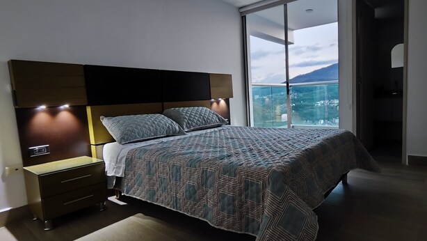 A modern bedroom with a large bed, a nightstand, and a built-in TV. The room has a large window with a city view.