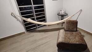 A room with a hammock, a cushioned chair, a small table with a decorative item, and a window with blinds.