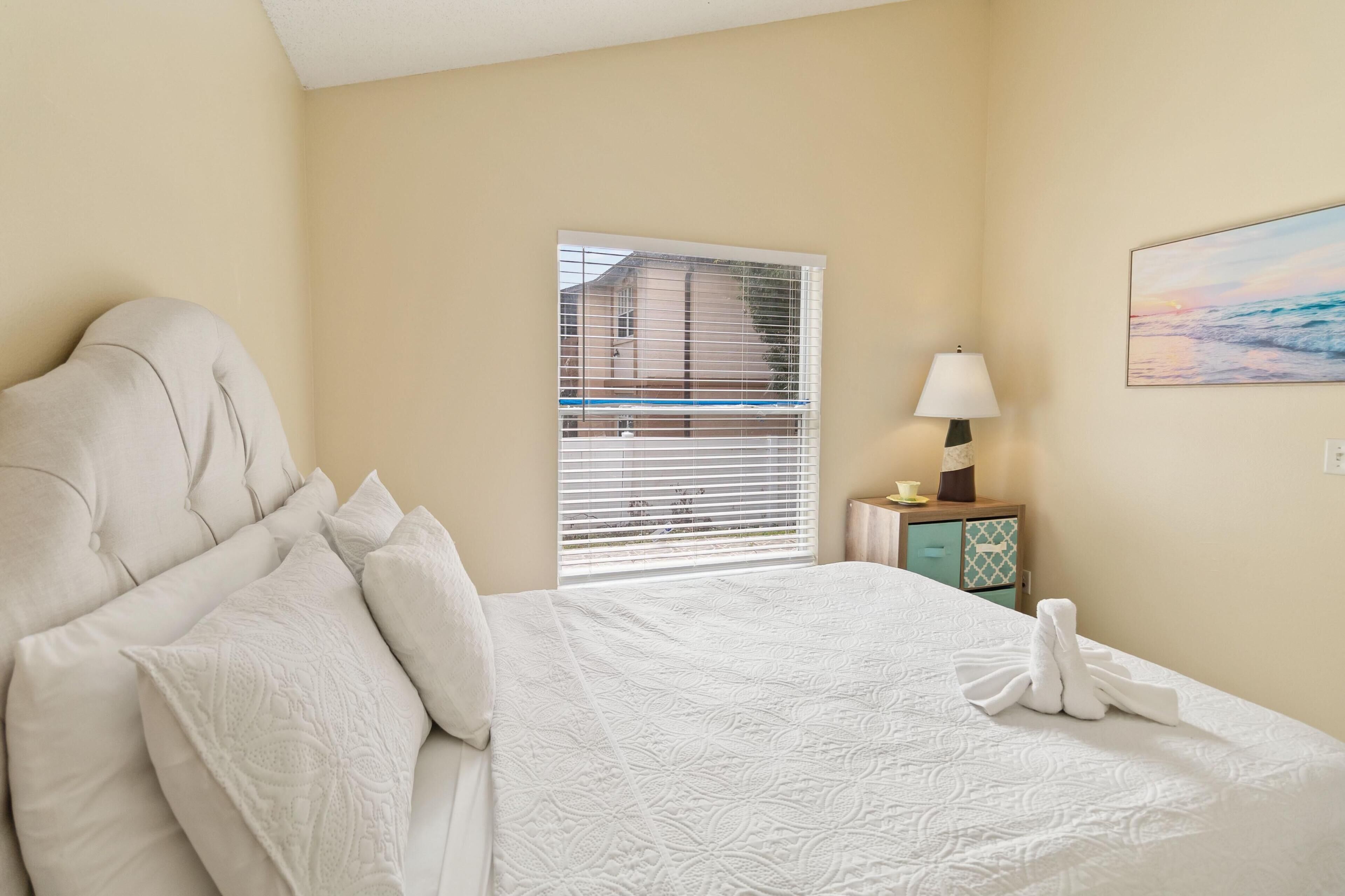 A neatly made bed with a white duvet, a bedside lamp, and a painting of a beach scene.