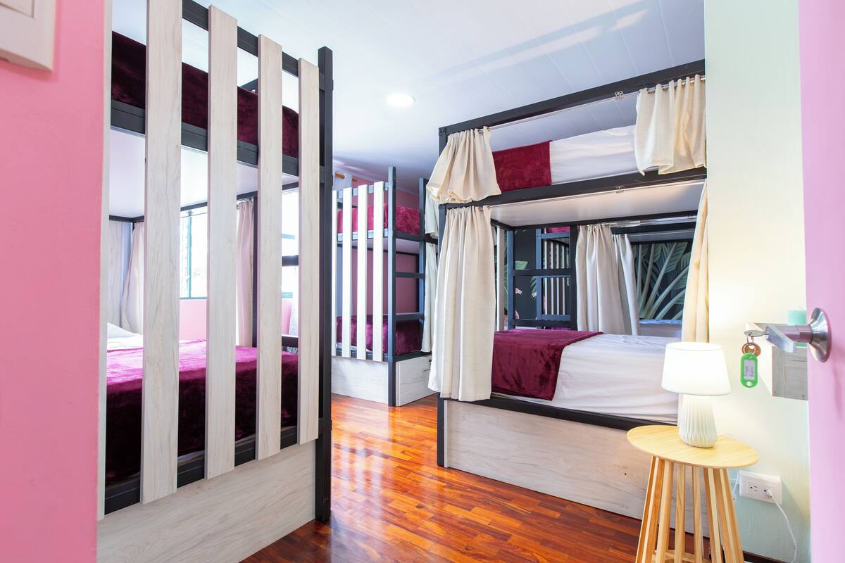 Een slaapkamer met een stapelbed, een houten vloer, een nachtkastje met lamp en een raam met gordijnen.