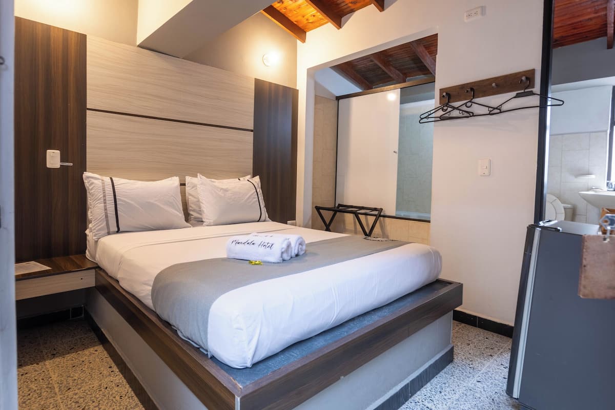 A modern bedroom with a bed, wooden ceiling beams, a small bedside table, a mirror, and a refrigerator.