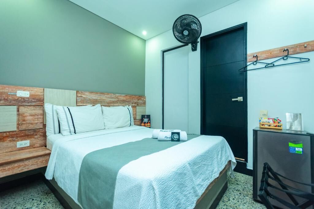 A hotel room with a bed, a wall-mounted fan, a door, and a small refrigerator.