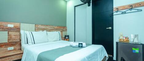 A hotel room with a bed, a wall-mounted fan, a door, and a small refrigerator.