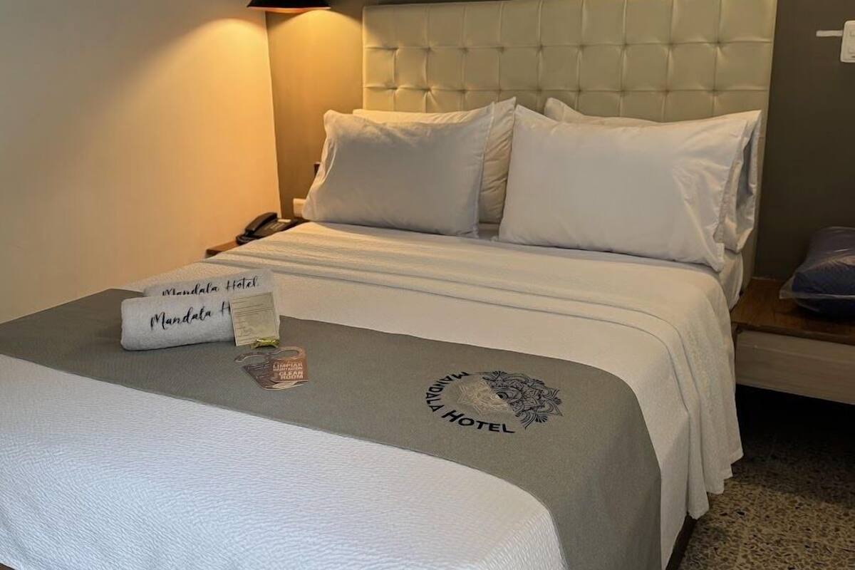 A neatly made bed with a white headboard and pillows, a bedside table with a lamp, and a sign indicating the hotel name.