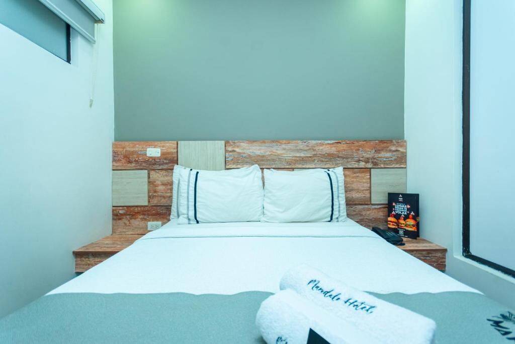 A hotel room with a bed, a headboard with a wooden texture, a bedside table with a phone, and a poster on the wall.