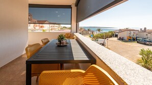 A balcony with a table and chairs, overlooking a coastal area with buildings and a clear sky.