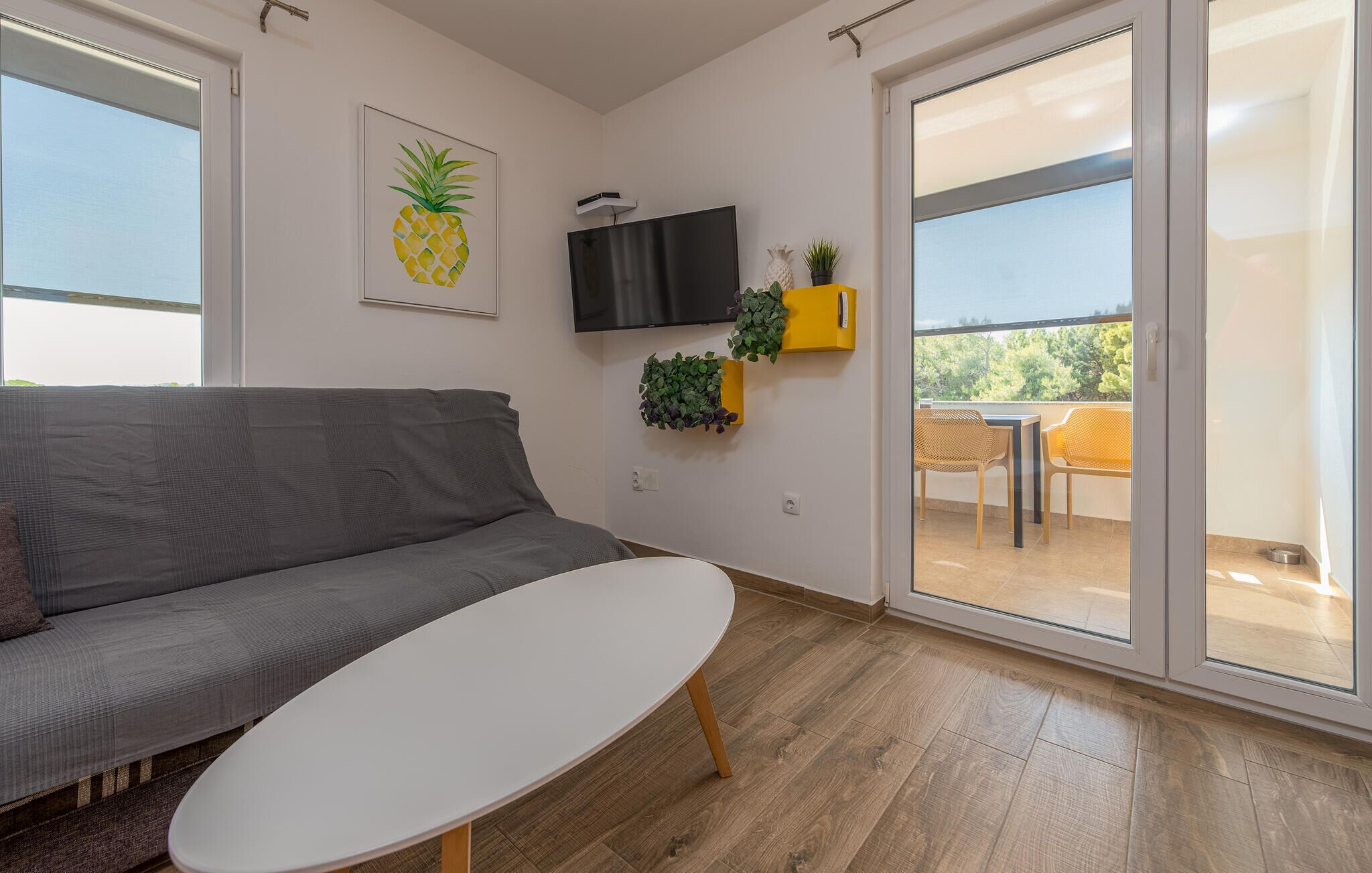 A modern living room with a grey sofa, a white coffee table, a flat-screen TV mounted on the wall, and a balcony with yellow chairs and potted plants.