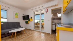 A modern living room with a grey sofa, a small white table, and a TV mounted on the wall. There are potted plants on a yellow shelf, and a sliding glass door leading to an outdoor area with a table and chairs.
