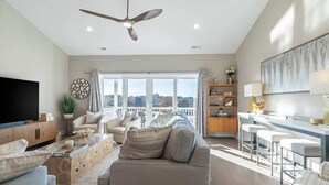 A spacious living room with a large TV, a ceiling fan, a sofa, a dining area with bar stools, and a view through large windows.