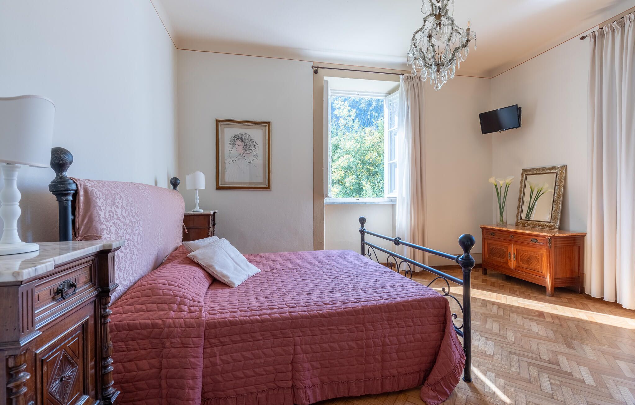 A bedroom with a bed, a dresser, a nightstand, a chandelier, a window, and a television.