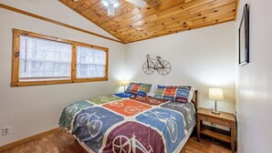 A bedroom with a wooden ceiling, a bed with a colorful bicycle-themed bedspread, two windows with blinds, a wall-mounted bicycle decoration, and a nightstand with a lamp.