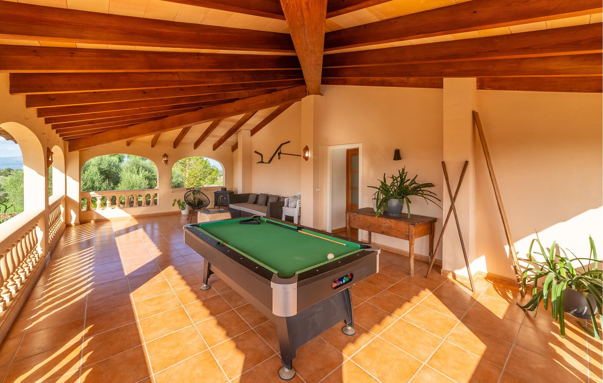 A pool table in a sunlit room with a balcony, wooden beams, and potted plants.