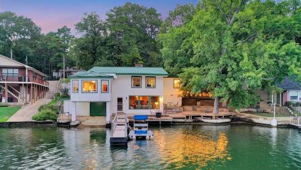 A houseboat docked at a pier with a green roof, surrounded by trees and a calm body of water. - Lake Haven (Hot Tub, Game Room) (Hot Springs)