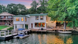 A waterfront property with a dock, a boat, and a house with a green roof and large windows. - Lake Haven (Hot Tub, Game Room) (Hot Springs)