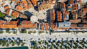 Vue aérienne d’une ville côtière avec des bâtiments aux toits de tuiles rouges, une place centrale et une zone de plage bordée de palmiers et de chaises longues.
