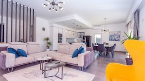 Un salon moderne avec un canapé, une table basse et un fauteuil jaune vif. On y trouve également un coin repas avec une table et des chaises, et une cuisine à l’arrière-plan.