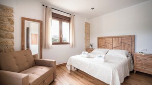 Una camera da letto con un letto, un comodino in legno, una sedia, un muro in pietra e una finestra con tende.
