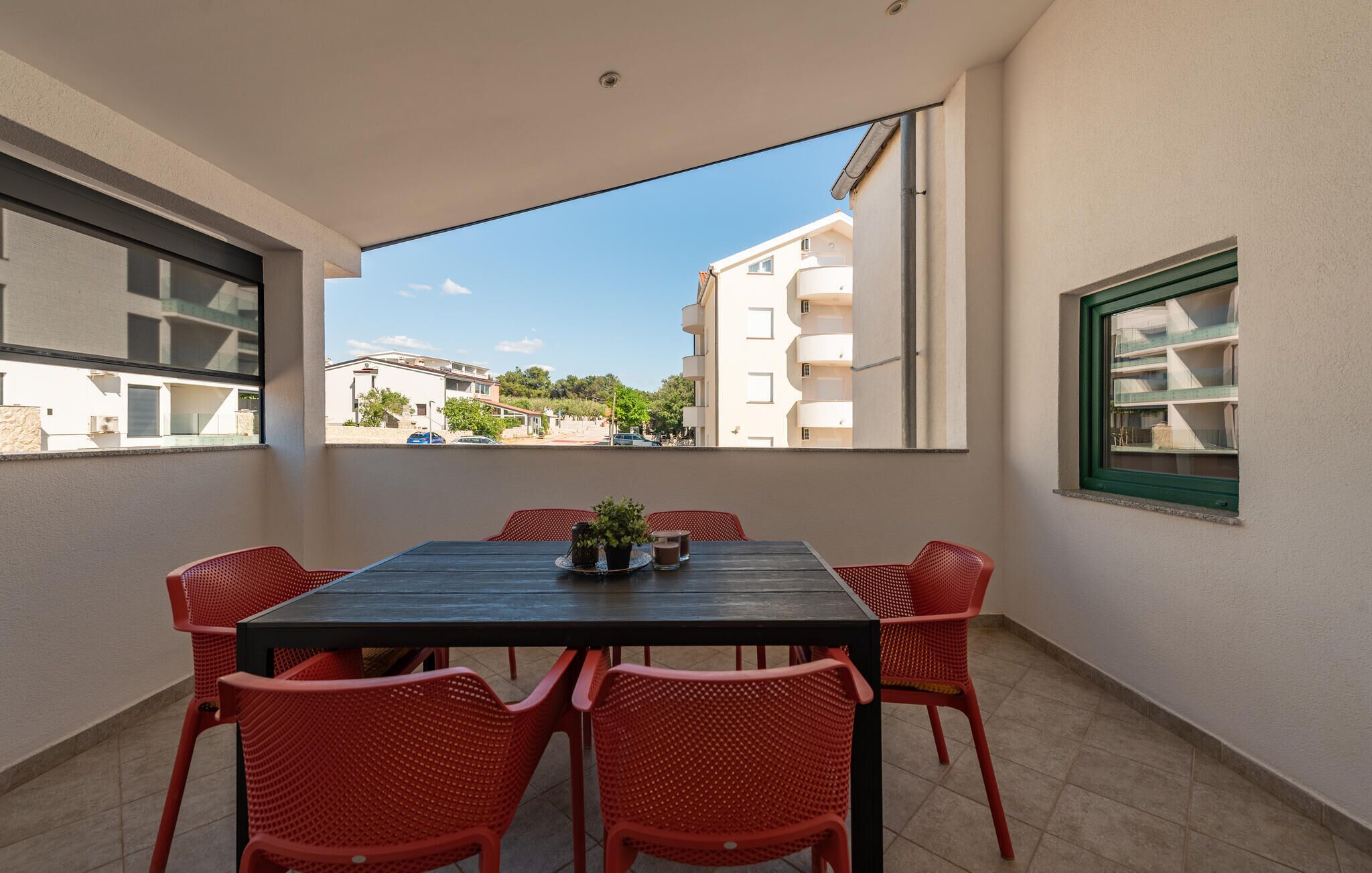 Un balcón con mesa y sillas, vista a edificios y una ventana con marco verde.