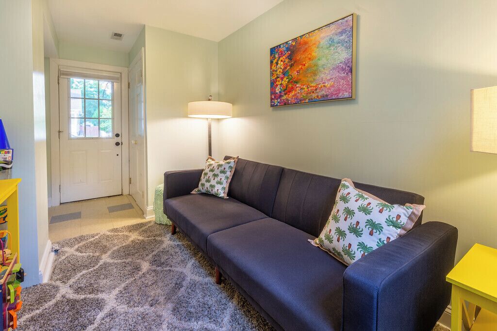 A living room with a dark sofa, two patterned pillows, a colorful abstract painting, a lamp, and a door with a window.