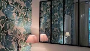 A room with a large mirror reflecting a bathroom with a shower and a toilet. The walls are covered with a tropical leaf pattern wallpaper. There is a small bedside table with a lamp.