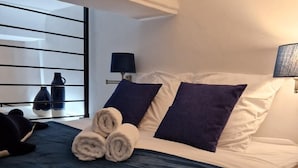 A neatly made bed with a dark blue bedspread, two pillows, and a rolled towel. There is a bedside table with a lamp and a vase.